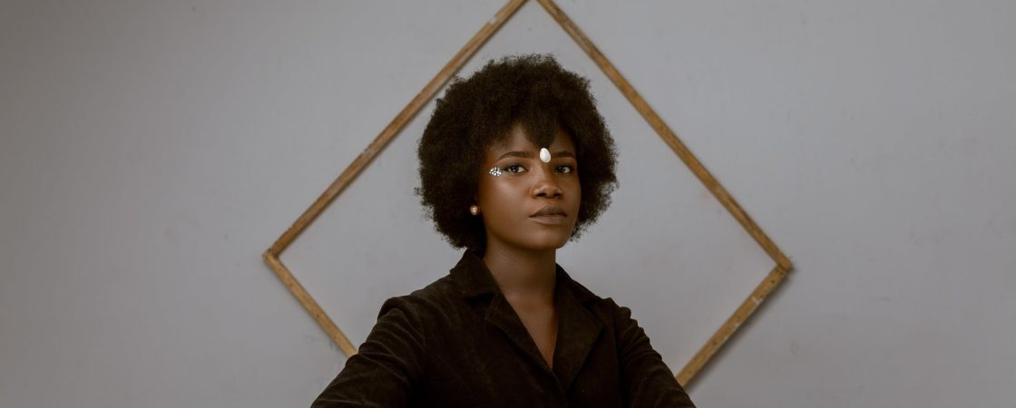 elegant young black woman with afro hair resting in room near decorated wall with frame