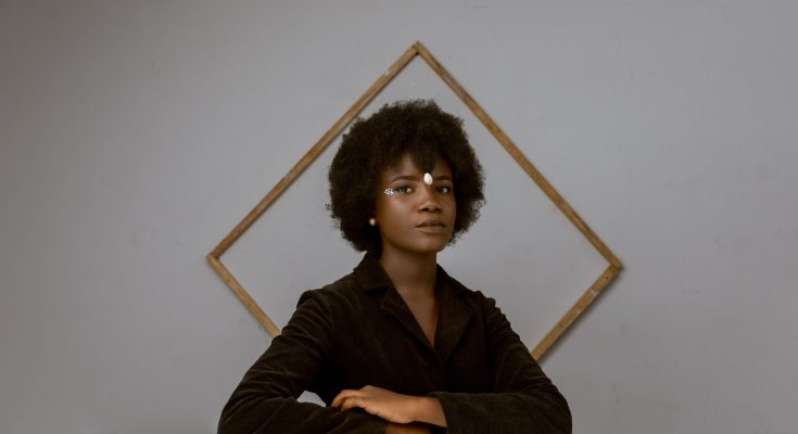 elegant young black woman with afro hair resting in room near decorated wall with frame