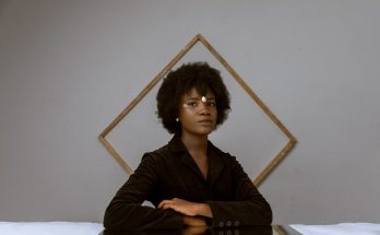 elegant young black woman with afro hair resting in room near decorated wall with frame